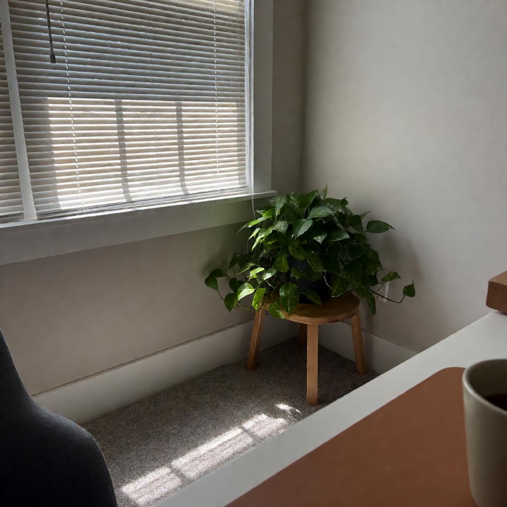 A slightly off-level smartphone photo of a home office corner with a healthy pothos plant on a low wooden stool, clean white baseboards, natural light from a nearby window, and a desk edge visible in the foreground.